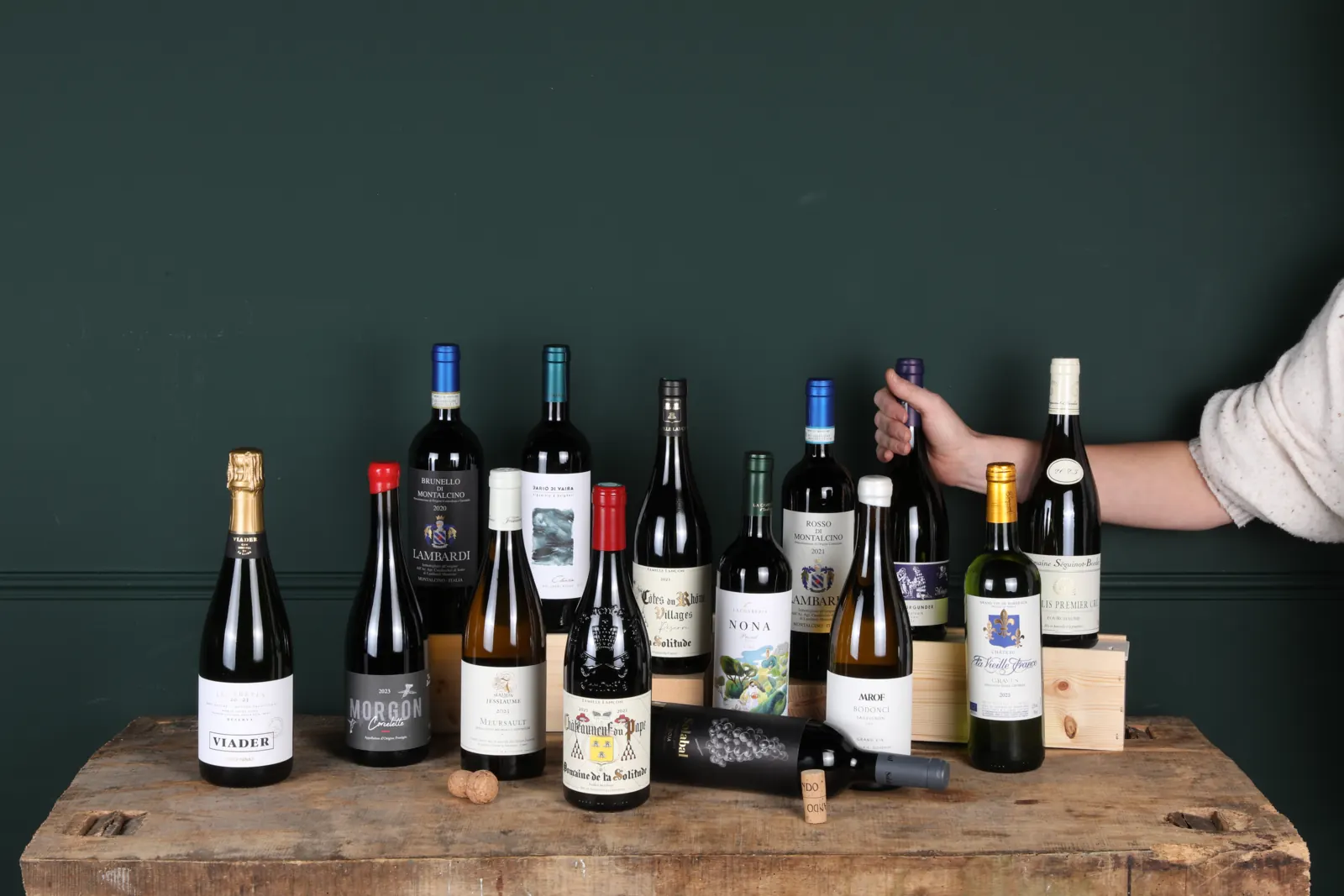 Wijnflessen op een houten tafel voor een donkergroene wand, een hand pakt er een uit