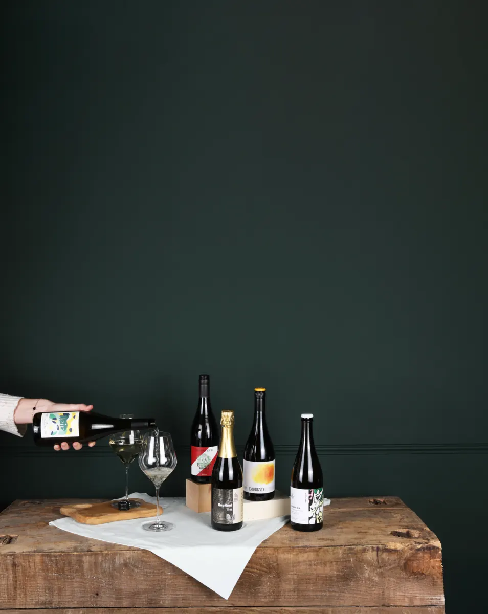 Wijnflessen op een tafel voor een donkergroene wand, een hand schenkt in