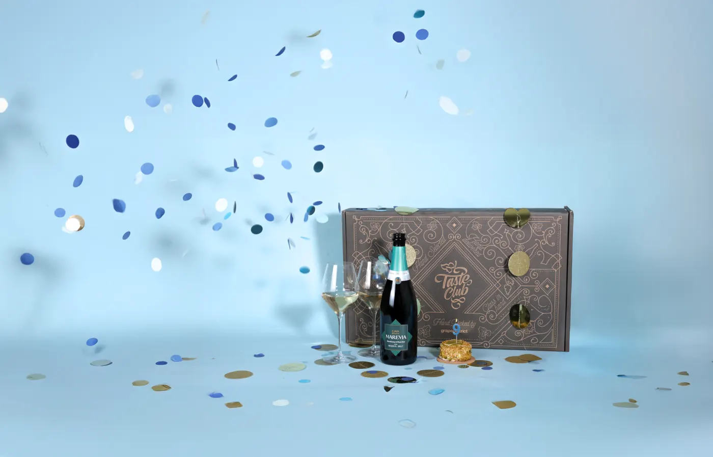 Taste Club-doos met confetti, een fles bubbels en twee glazen tegen een lichtblauwe achtergrond