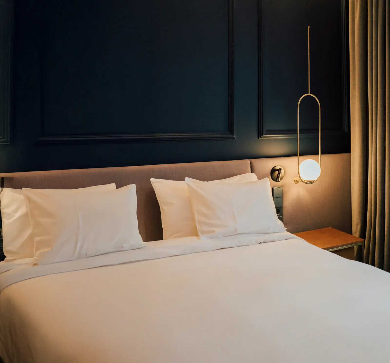 Hotelkamer met diepblauwe lambrisering, wit beddengoed en een slanke hanglamp